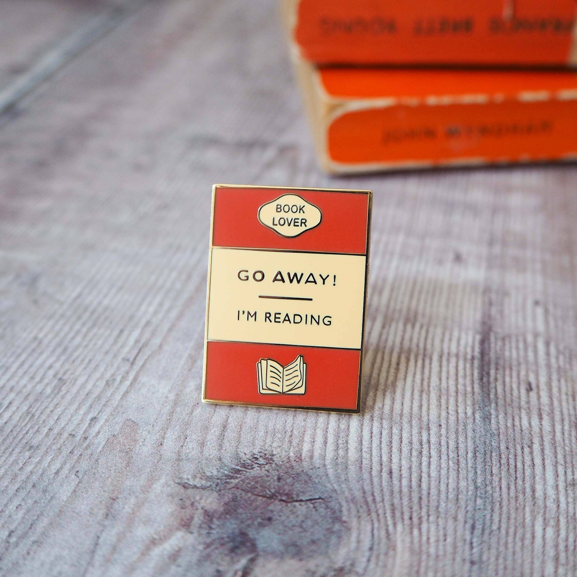 Close up photo of "Go Away I'm Reading Enamel Pin" on a wooden bench.